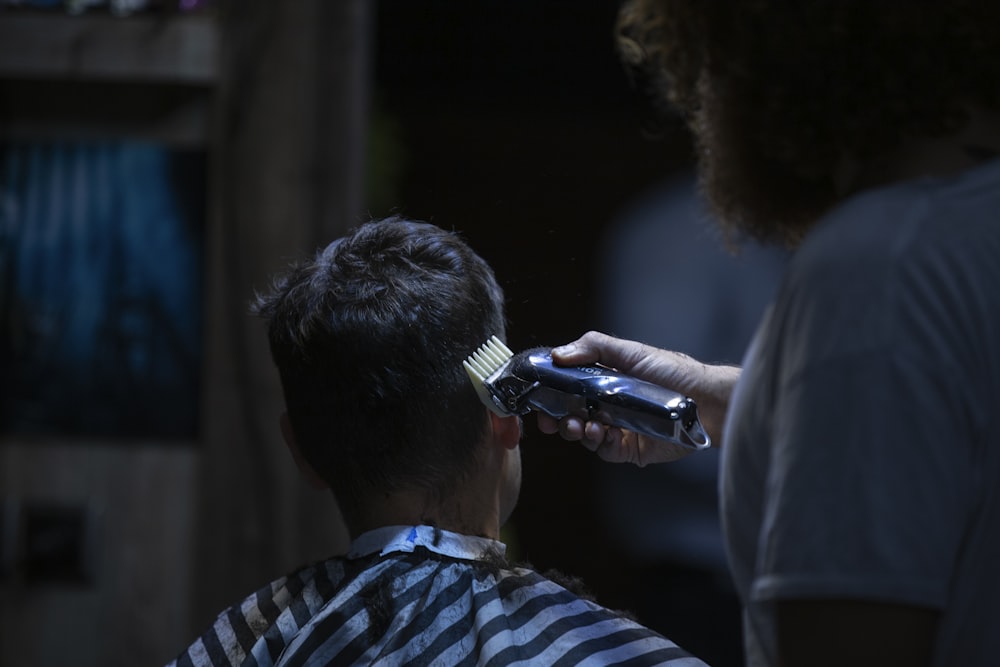 A bearded person holding a clipper cuts the hair of a person wearing a striped cape