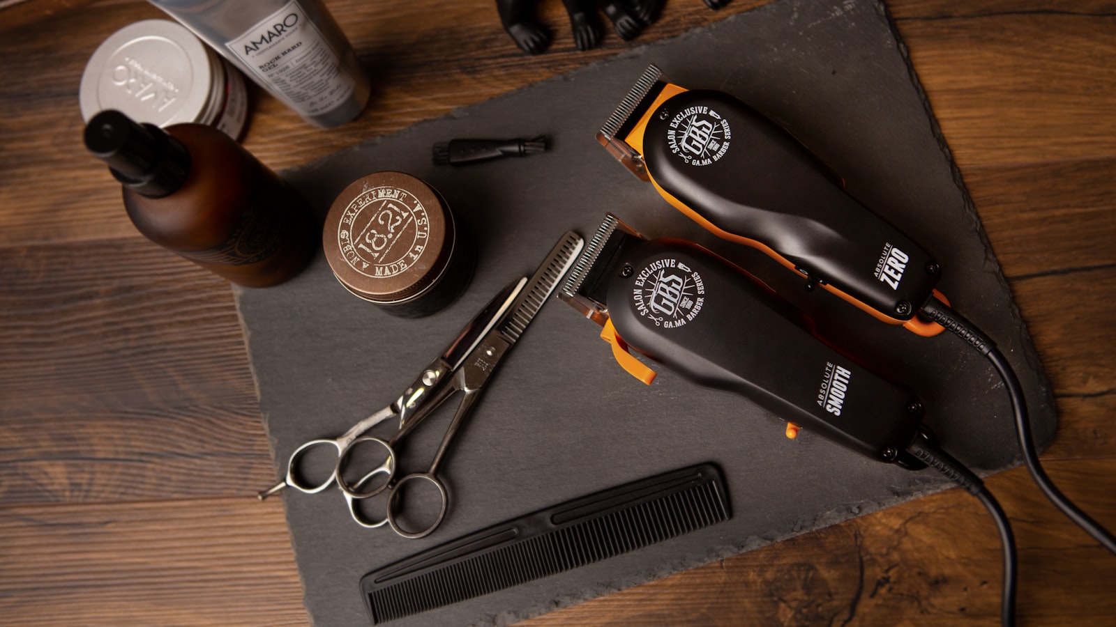 Two hair clippers, scissors, a comb, and grooming products rest on a slate board